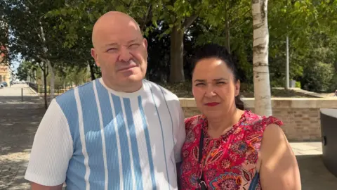A man and a woman stand together on a pavement in front of a public garden. They have serious expressions on their faces. The man has a shaved head and wears a T-shirt with blue and white vertical stripes. The woman on the right has black hair, tied back and wears a red, pattered top.