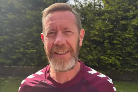 Colin Brown Colin Brown has short fair hair and smiles at the camera. He wears a maroon Hearts FC strip and is standing in a garden with a bush behind him