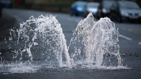 Water spurts upwards out of a drain on a road.
