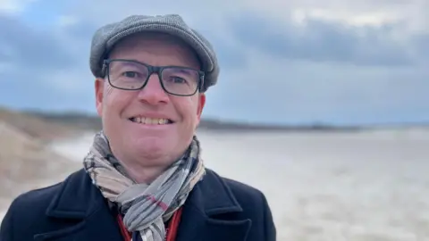 Stuart is standing on a sandy beach wearing a dark coat, a patterned scarf, and a flat cap. The background shows a cloudy sky and distant shoreline. 