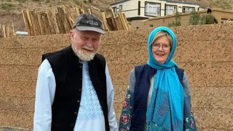 PA Media Peter y Barbie Reynolds sonríen a la cámara frente a una pared de ladrillo naranja. Peter es un anciano que usa una parte superior azul de manga larga, un chaleco negro y una gorra gris, mientras que Barbie usa un pañuelo azul brillante, una chalea azul marino y una tapa azul estampada.