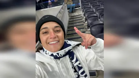 Mandy Damari Emily Damari, a young woman wearing a Spurs scarf and a black beanie hat, smiles in the stands of a football ground