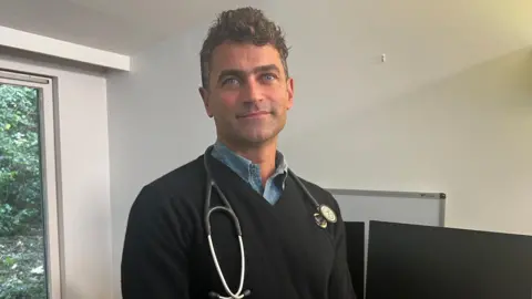 Dr Tom Sutherland is looking straight at the camera. He is wearing a dark blue jumper and has a stethoscope round his neck. His has blue eyes and short curly dark hair. 