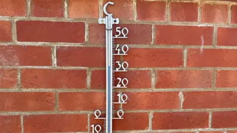 BBC Weather Watchers/Suzies View A silver coloured thermometer fixed to a red brick wall. It has C at the top and to its right are numbers ranging from 0 to 50. The blue temperature gauge appears to have reached 33C.