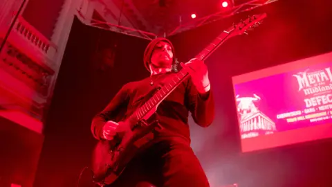 A guitarist on stage wearing a beanie hat. He is illuminated by red stage lighting