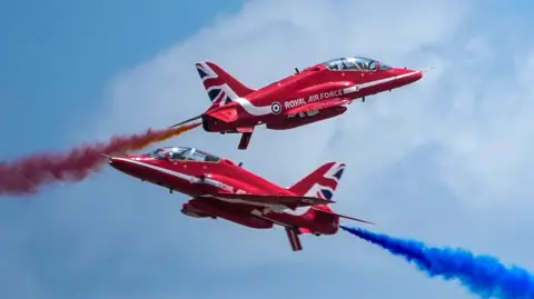 Two Red Arrow planes flying diagonally upwards in opposite directions. They are marked with a white Royal Air Force sign and a Union jack. The plane flying to the right has a trail of red smoke while the one flying to the left has a trail of blue smoke