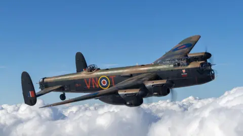 A bomber plane with black sides and camouflage patterns on its roof, flying through a blue sky with some white clouds. Letters on the side read VNOT