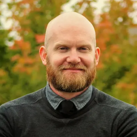 Jenny Routledge A man with shaved head and ginger beard smiles for a portrait style shot against a background of red-green autumnal leaves.