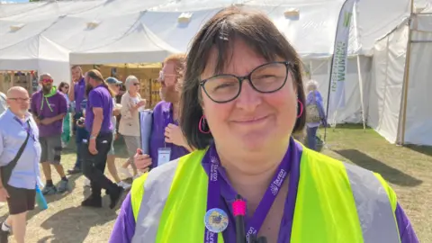 Beccy Fox, is standing outside a white marquee. She is wearing a purple shirt and yellow high-vis vest.