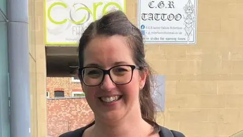A young woman with tied back long brown hair and round black glasses smiles at the camera