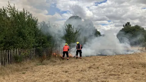 JOANNA TAYLOR/BBC Two fire fighters in orange and black hi-vis uniforms with yellow helmets are in action pointing hoses and beaters at a fire behind a wooden gate enclosing trees. Next to them is a large patch of burnt grass with smoke rising from it. 