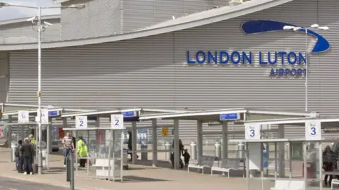 London Luton Airport Operations The exterior of London Luton Airport. The branding on the airport building is visible, in blue, and in the foreground are bus stands and some people standing in the sunshine.