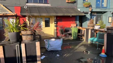 BBC The outside of The Vault premises. There is an industrial bag outside of the entrance and there is blocks of wood with plant pots on top of it creating an area in front of the entrance. 