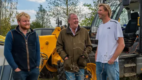 Prime Video Andy Cato laughing with Jeremy Clarkson and Kaleb Cooper on Diddly Squat farm in the Cotswolds, standing in front of a telehandler.