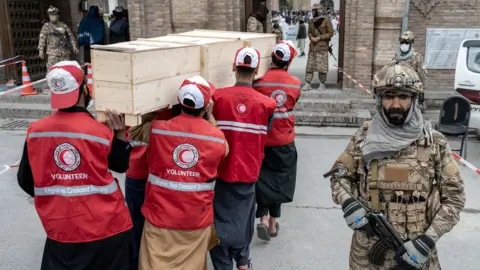 Taliban security personnel stand guard as Afghan Red Crescent Society volunteers carry the coffins of victims who were killed by a Pakistani airstrike that struck a drug rehabilitation center, during the second mass funeral ceremony outside the Eid Gah Mosque in Kabul on March 26, 2026.