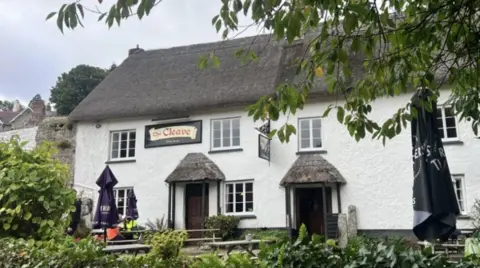 Outside The Cleave Inn in Lustleigh. The property is white with a thatched roof. Benches are outside the pub.