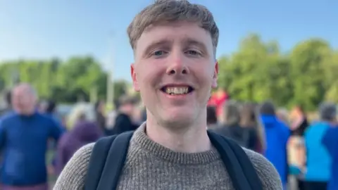 A man smiles into the camera. He has fair hair and is wearing a grey jumper. He has black backpack straps over his shoulders.