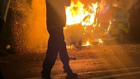 Police van on fire with a person standing in front of it
