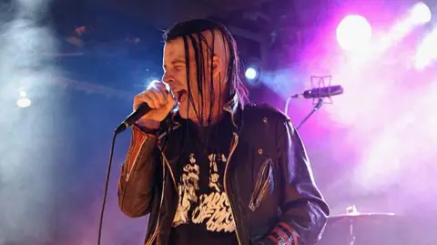 A man with semi-shaved head and black braids sings into a microphone. There are stage lights shining through mist on the stage behind him.