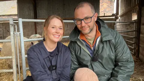 Laura and Tim are sat in front of their herd of sheep. Laura has dark blonde hair, tied back in a ponytail and wears a blue overall. Tim has a green jacket on over a blue top and wears blue-rimmed glasses.
