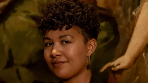 A young woman with curly short hair smiles at the camera. 