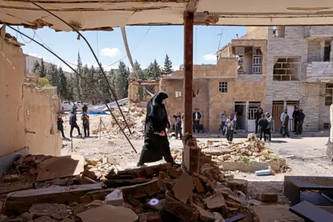 AFP via Getty Images A woman walks past the residential buildings that were damaged by recent strikes at Vahdat town in Karaj, southwest of Tehran on 3 April, with bystanders and other buildings in the background against a sunny blue sky.