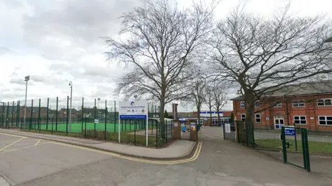 Green fencing surrounding a multi-sports pitch and an entrance sign for Ibstock School in front of trees without leaves and a red-brick building