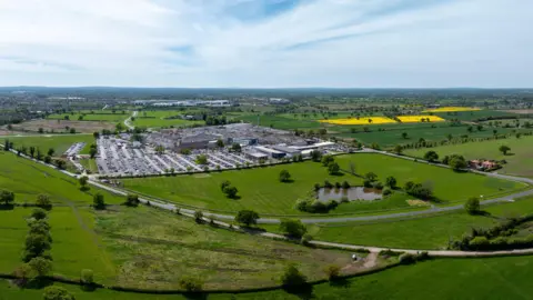An aerial view of the Leighton Hospital site. A large car park is visible on the left with a collection of buildings alongside. The site is surrounded by fields and roads can be seen running around the site.
