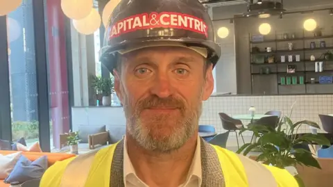 Paul Richards is wearing a black hard hat, and a high vis jacket over a blazer. He has a greying beard.
