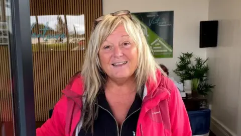 Jackie Leek smiling opening a door wearing a black top and salmon-coloured jacket