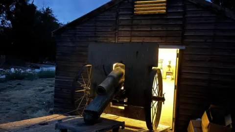 Great War Huts A rusty short-barrelled 10.5cm howitzer gun with its nose down on a wooden pallet. Behind it is a wooden hut and a large open door, from which light is glowing.