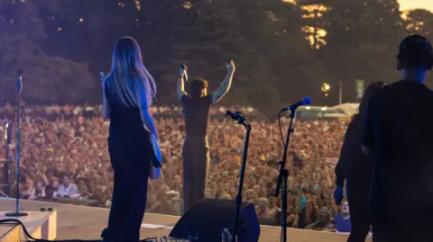 Cuffe and Taylor Tom Grennan on stage. The view is of his back with the crowd in front. He has his arms up. A female guitarist is on stage with him.
