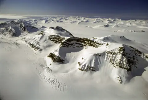 Getty Images - Doug Allan Aerial view of rugged, snow-covered mountains and ridges rising from a vast polar ice sheet, with layered rock faces visible beneath the white snow and ice.