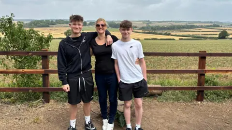 Emma Parker Emma wearing a black t-shirt and trousers smiling in between her two sons. Her elder son, on the left, has his arm around her and is wearing casual attire. her younger son is on her right, wearing casual attire. They are standing in front of a wooden fence, with fields behind it. 