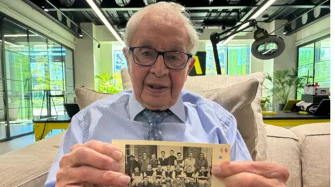 Griff Bowen poses for a photo on a brown sofa in a large, light-filled meeting room. He has white hair and black-rimmed glasses, and wears a striped blue shirt and patterned dark-blue tie. He is holding up an old photograph of a football team from 1948, where he can be seen on the right hand side with longer black hair. 