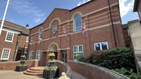 Exterior of South Kesteven District Council offices. It is a two-storey brick building with the council's coat of arms over the main door and four flags mounted on poles on the front of the building