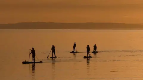 Jean Penman Five paddleboarders glide across calm water at sunset, appearing as dark silhouettes against a warm golden sky. Distant hills stretch across the horizon, reflected in the still sea.