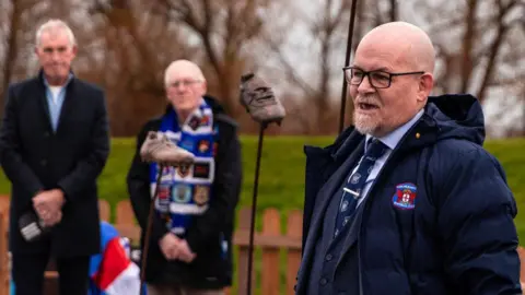Carlisle United Club chaplain Dave Allen standing in front of the statues. He is wearing a suit, club tie and a large blue coat with the club's badge on its breast. He has a grey beard and bald head. Three boots are displayed on large poles behind him. Two people stand in the background with their hands held in front of them in respect.