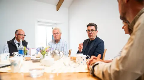 PA Media National Farmers Union President Tom Bradshaw (left) and Prime Minister Andy Burnham (middle write) sat at a table. There are other people around them. There are glasses and mugs and a teapot on the table.