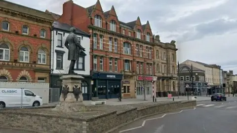 A view of a statue on Church Street, Hartlepool, with shops and parked cars along the street.