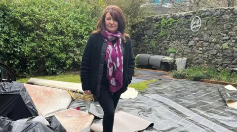 Sophie Ingram Sophie is standing in a garden with a hedge behind her and a stone wall to her right. She is standing on damaged linoleum floors which have been removed from her shop due to storm damage. She is looking at the camera and has long brown hair. She has a purple scarf, a black jacket and black skinny jeans.