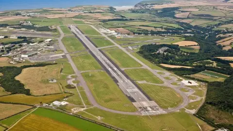 Aerial view of a plane runway. It is surrounded by green fields. The sea is in the background. 