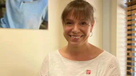 Fiona está sentada en una silla dentro de casa, sonriendo a la cámara, con una camiseta blanca con una insignia de "Jersey Child Care Trust", y una foto borrosa de un niño y un adulto en la pared detrás.