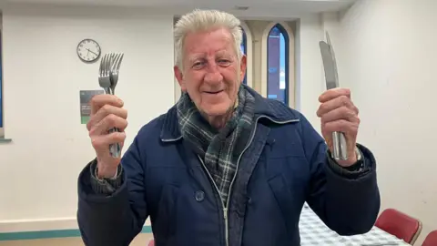 BBC/ Elizabeth Baines Bernard Clark, smiling with knives and forks in his hands.