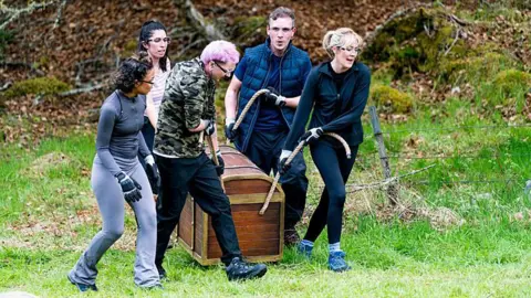 BBC/STUDIO LAMBERT Alex, (centre with pink hair) and four of his fellow contestants, carry a chest in one of the challenges - they are wearing trainers and safety goggles