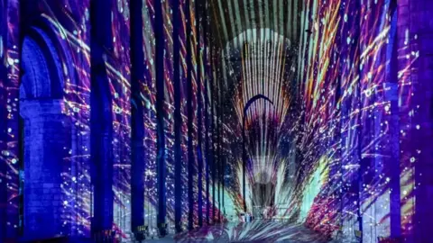 The inside of Ely Cathedral is covered with lights which are projecting on to the building and are showing multiple colours. 