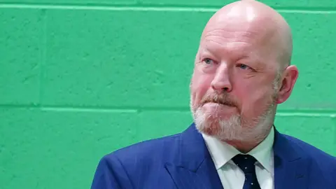 PA Media Simon Danczuk Reform UK candidate who is bald with a white beard wearing a blue suit with blue and white spotted tie. He stands in front of a green wall.