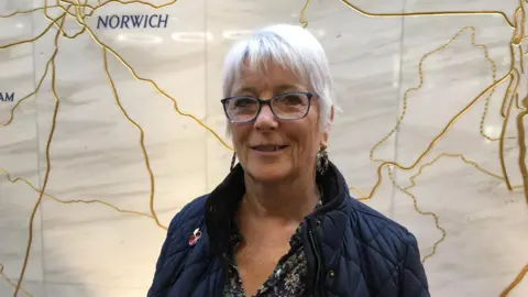 JO THEWLIS/BBC Alison Thomas is wearing a dark blue coat over a dark, patterned top. She is also wearing glasses and standing front of a marble wall.
