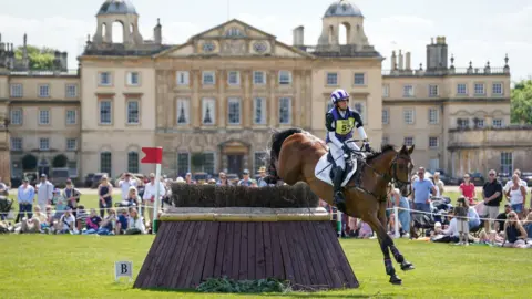 PA Media A horse and rider clear a jump obstacle in front of the main house at the Badminton Estate in Gloucestershire. In the background several spectators can be seen watching on