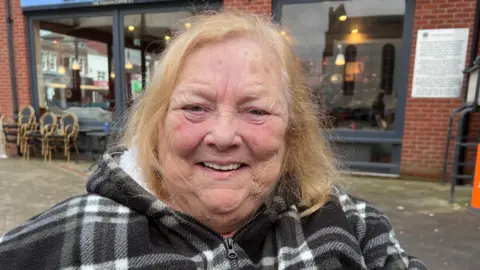 Marie has red shoulder-length hair and is wearing a checkered black and white coat.